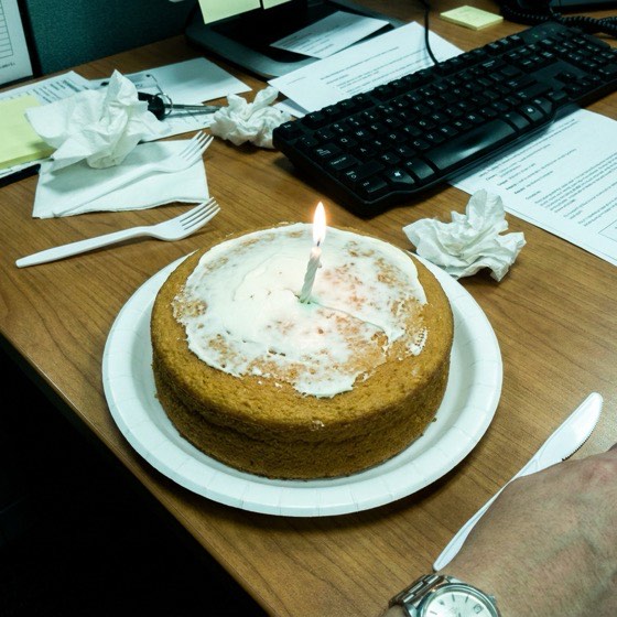 A supermarket sponge cake with one lit candle, on a cluttered office desk.