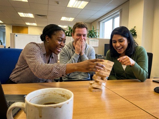 Three colleagues laughing at a tower of wooden blocks about to topple on a meeting room table.