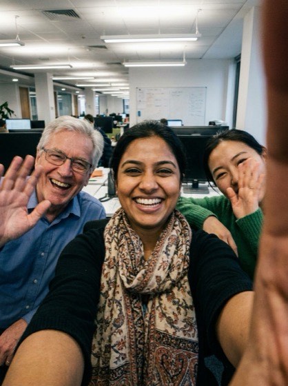 Three colleagues crowded into a selfie in an open plan office, waving at the camera.
